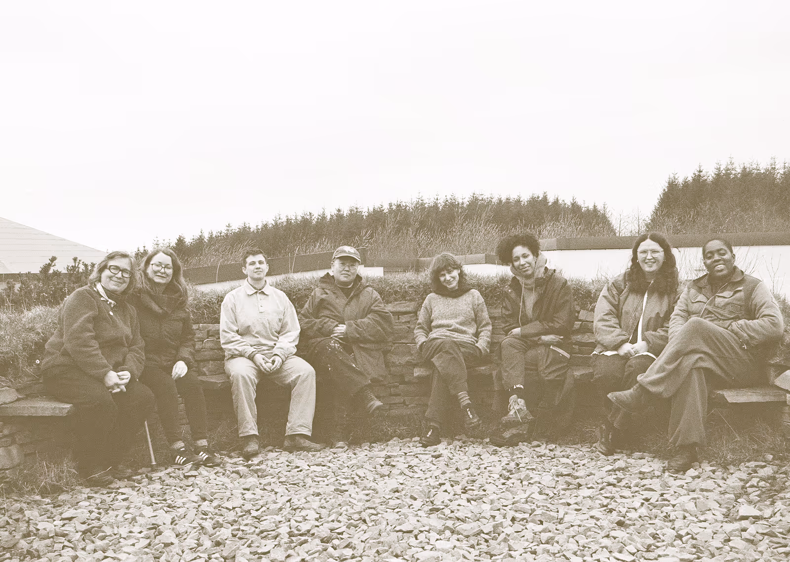 A landscape image with a goldy brown filter across the photograph. A group of eight people sit in a semi-circle on slate seats sunk into a slate wall. The top of the wall is grass covered and the ground at their feet is covered in flinted slate a slightly lighter shade than that of the wall. The group members are all smiling towards the camera, some leaning into each other and all are wearing woollen jumpers, coats and scarves and the sky is a clouded bank in the background. Behind them are low buildings with white walls, most with flat roofs except one slanted to the left side. The tops of conifer trees are in the distance. The group in order from left to right is made up of Deirdre O'Mahony, Bridget O'Gorman, D. Mortimer, Taey Iohe, Hannah Wallis, Libita Sibungu, Iarlaith Ní Fheorais and Onyeka Igwe during the Deep Time slow retreat at Common Knowledge, Co. Clare.