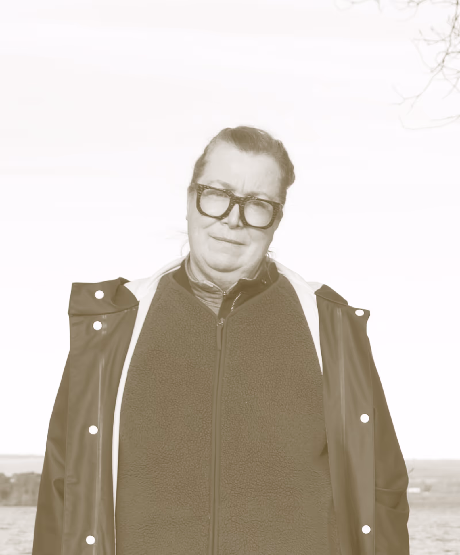 A portrait image with a goldy brown filter, of a person from the shoulders up facing the camera with a neutral expression. They wear thick rimmed glasses and have their hair up, wearing a fleece.