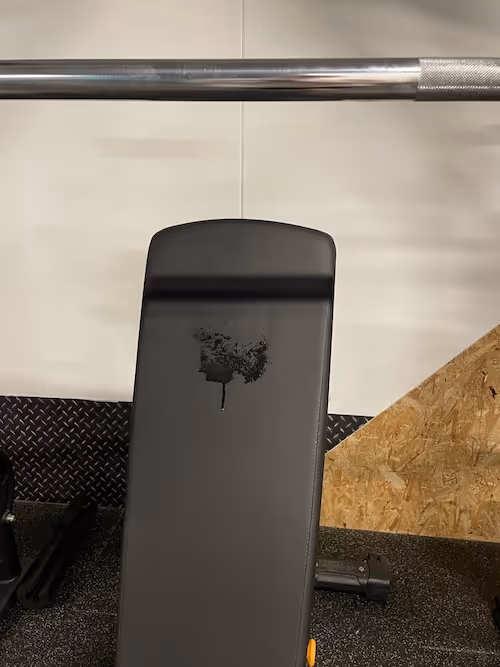 A steel bar cuts across the top of a portrait image, casting a shadow at the top of a back rest on a piece of gym equipment. A sweat patch drips down the back rest which is against a speckled tiled floor, a corner of woodchip against the wall and a strip of metal protection at the bottom of the wall before becoming plain white walls. The start of another piece of gym kit peeks in at  the left edge of the image. 