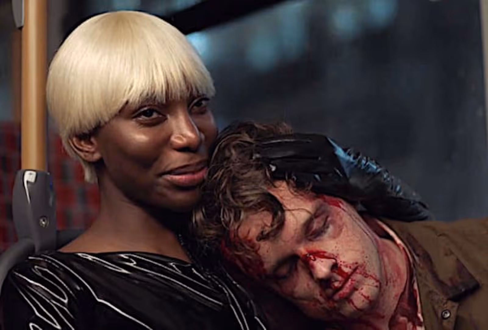 A landscape image of two figures from the shoulders up, sitting on a bus. The figure on the left is wearing black latex and has short pixie style blonde hair. They are smiling forwards and are cupping the head of another figure onto their shoulder through black gloves. The second figure has curly brown hair, is wearing a khaki jacket and their face is covered in blood. They have their eyes closed, leaning their head into the shoulder of the first figure. In the background is a blur of buildings through the window of the bus.