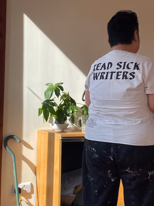 A portrait image of a figure from the knees up, with dark cropped hair facing away from the camera wearing a white t-shirt that says “read sick writers’ in black serif cursive and paint spattered trousers. To the left of the figure sun slants in on the pale walls across a wooden sideboard and a pale green walking cane leaning against the wall. There is a leafy green plant on the sideboard and the edge of a glass mug disappearing behind the figure. 