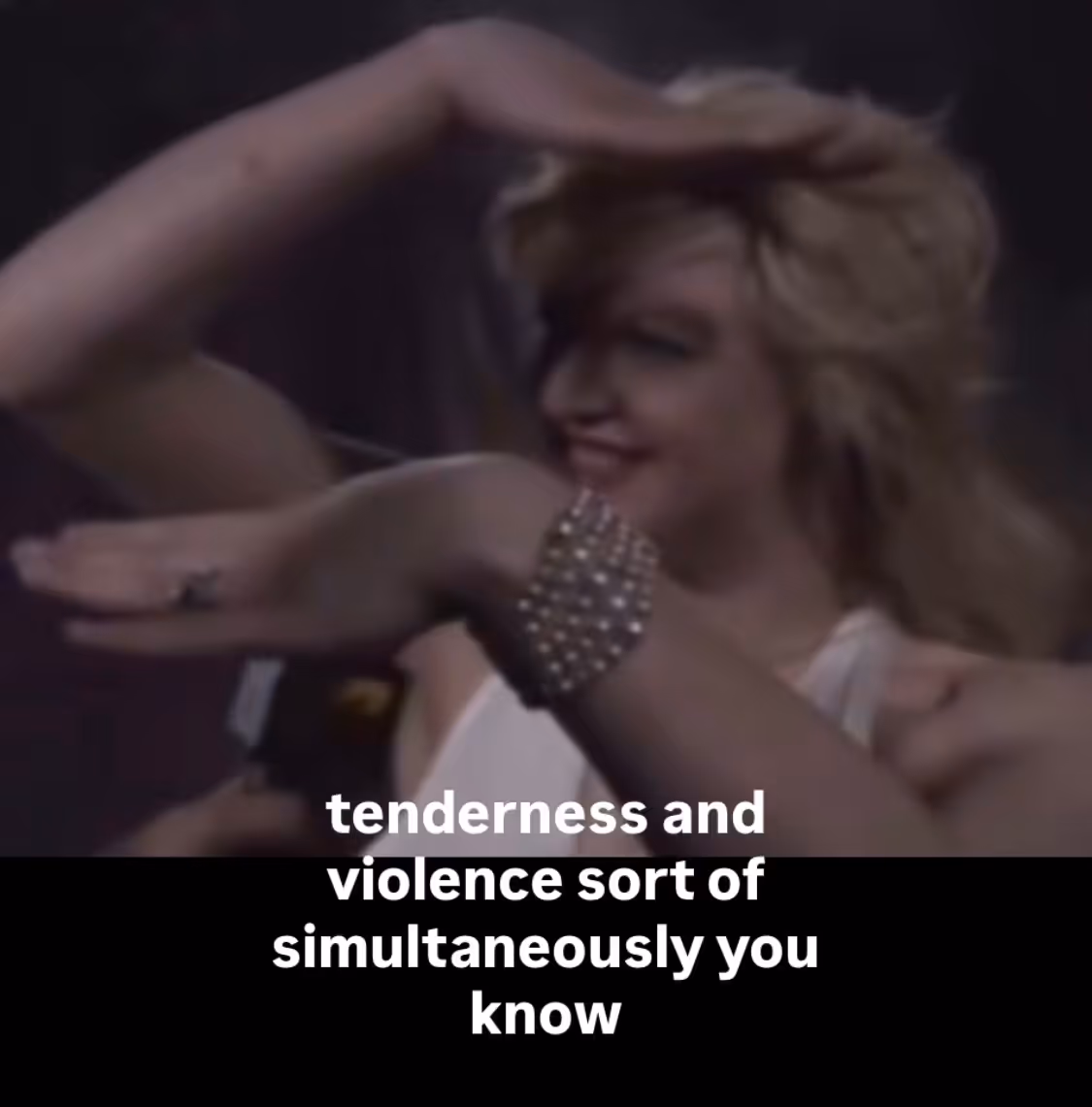 A square image, the top two thirds a figure with blonde hair smiling at someone holding a red microphone towards them, that reads MTV on the side. The figure is holding their right arm up in show of strength or celebration. The lower third of the image is plain black and written in white text across both sections, starting just below the microphone it reads, “tenderness and violence sort of simultaneously you know”. The figure is Courtney Love.